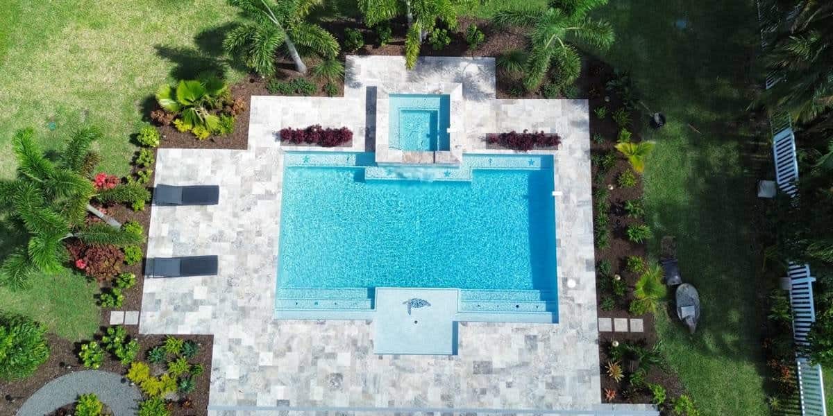 DIY pool paver installation (1) Aerial view of a custom swimming pool with light-colored stone pavers surrounding the deck, showcasing professional DIY pool paver installation with tropical landscaping, lounge chairs, and a decorative spa area with integrated water features
