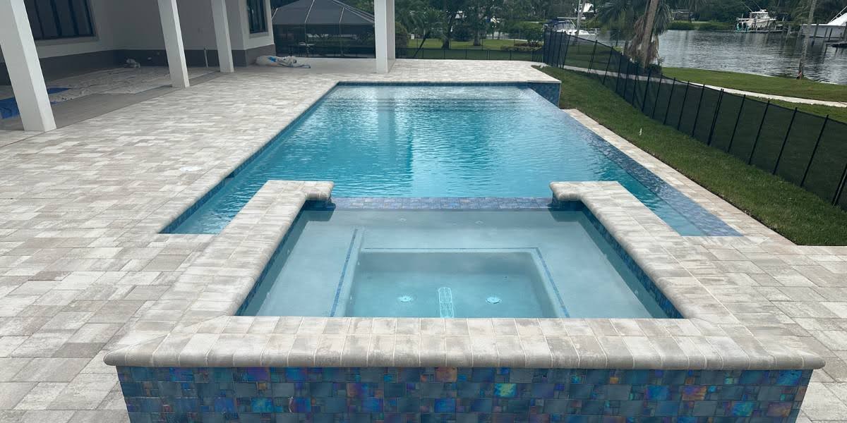 maintain pool pavers Luxurious backyard pool with travertine pavers surrounding the deck, hot tub with decorative blue tile, and water view - a perfect example of well-maintained pool pavers that enhance outdoor living spaces