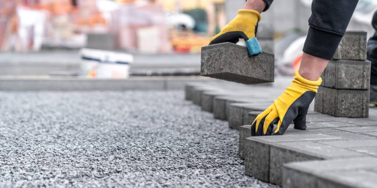 Worker placing pavers on gravel foundation while wearing gloves, showing how to pave a driveway using interlocking blocks.